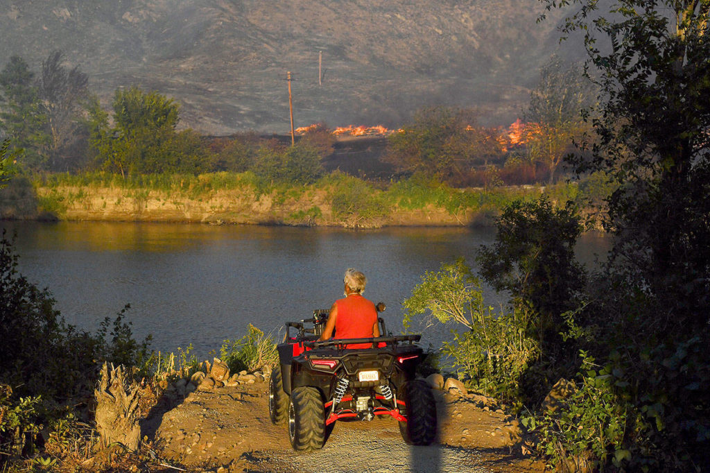 Mike Menalia pauses on his ATV on the banks of the Okanogan River and watches a pile of old railroad ties burn Monday in Okanogan. The river stopped the Cold Springs Canyon Pearl Hill Fire from crossing and burning his home. (Tyler Tjomsland/The Spokesman-Review via AP) 

