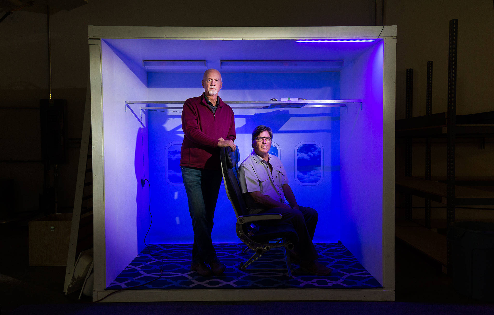 Under UV light, CEO Robert Olson (right) and President Gregory Strand sit in a mockup of an airplane interior on Sept. 14 in Everett. Waypoint Aeronautical, an Everett aerospace company, is developing an ultraviolet (UV) system to disinfect planes, trains and buses. (Andy Bronson / The Herald)