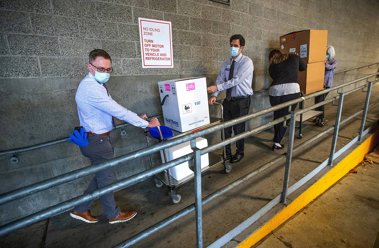 University of Washington Medical Center Montlake campus pharmacy administration resident Derek Pohlmeyer (left) and UWMC pharmacy director Michael Alwan transport a box containing Pfizer-BioNTech COVID-19 vaccines toward a waiting vehicle headed to the UW Medical Center’s other hospital campuses on Monday in Seattle. (Mike Siegel/The Seattle Times via AP, Pool)