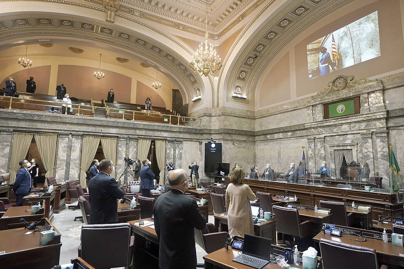 Senate members stand as a ceremonial presentation of colors is done virtually on a video screen above Monday, Jan. 11, 2021, at the Capitol in Olympia, Wash. Washington state's Legislature convened Monday under a large security presence because of concerns about efforts by armed groups who might try to disrupt the proceedings or occupy the Capitol, which is closed to the public due to the ongoing pandemic. (AP Photo/Ted S. Warren)