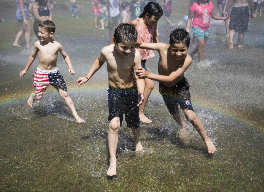 David Parada, 7, left, and Abel Parada, 8, run through a heavy spray of water creating a rainbow Saturday at Walter E. Hall Park in Everett. The Everett Fire Department set up a fire hose sprinkler station to help people cool down and escape the heat. (Olivia Vanni / The Herald)
