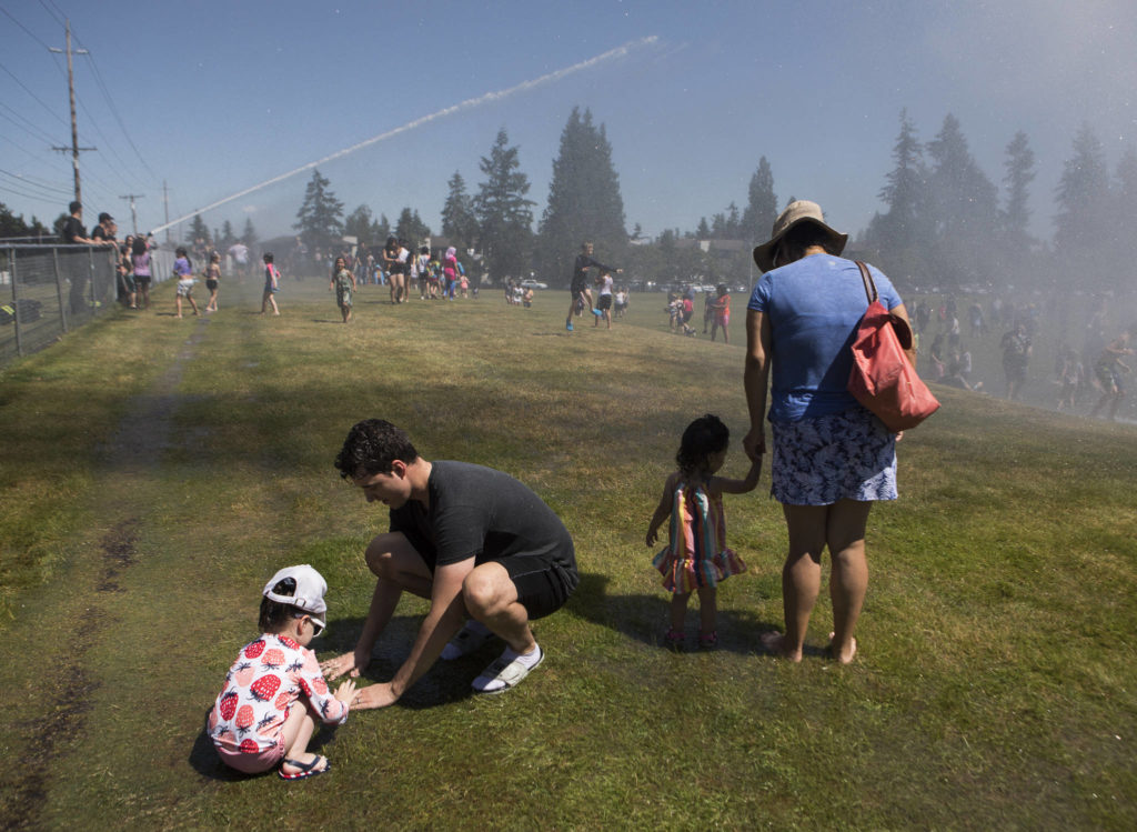 Derek Bock and his daughter Olivia, 3, left, play in a small puddle Saturday at the Everett Fire Department’s fire hose sprinkler station at Walter E. Hall Park in Everett. (Olivia Vanni / The Herald)
