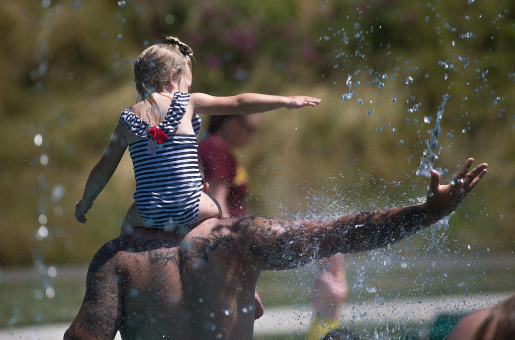 Keeping cool in record setting heat, Beau Jess and daughter River, 3, reach for falling water as they play at the Splash Pad in Haller Park on Monday in Arlington. (Andy Bronson / The Herald)
