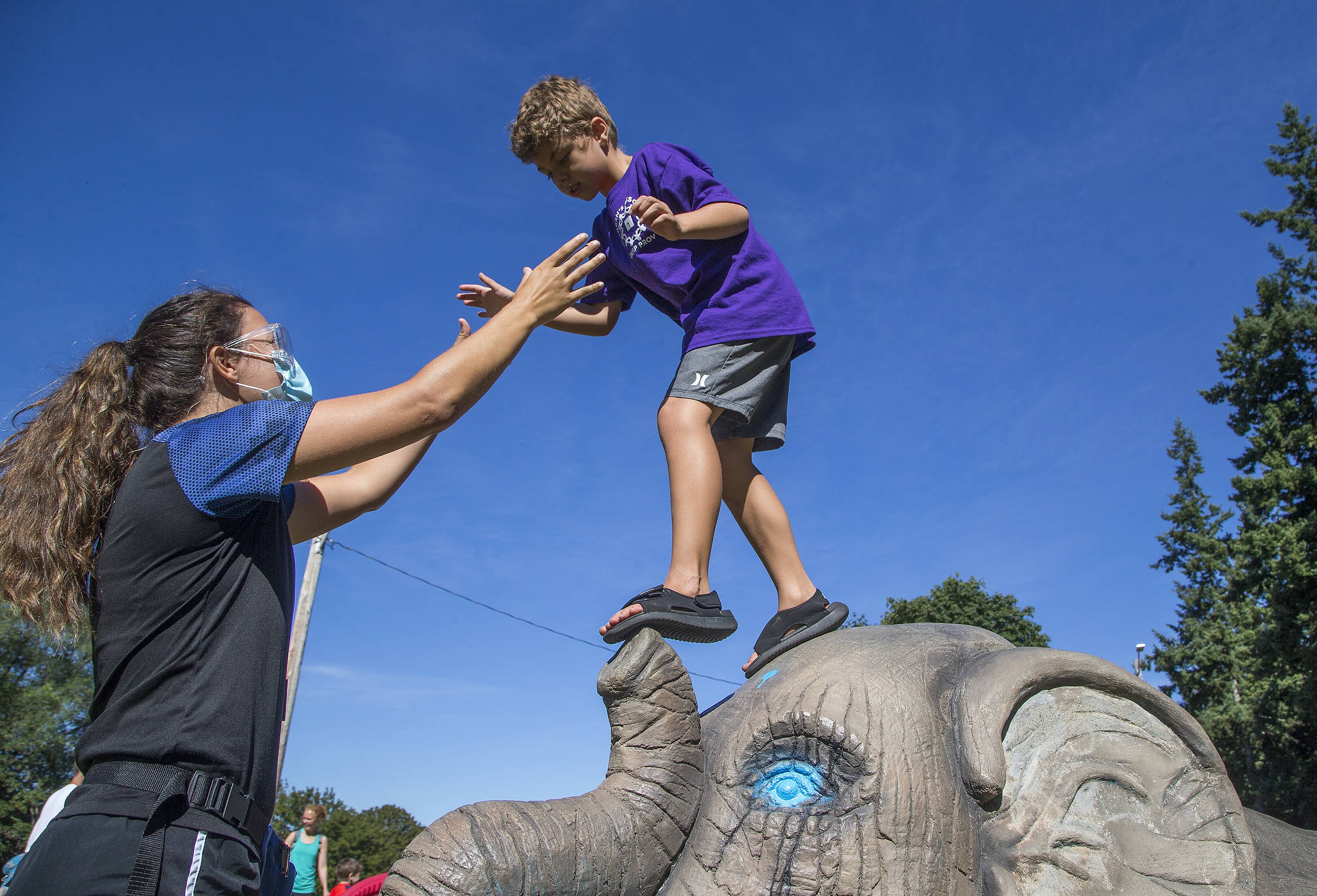Haaken Williams, 6, balances on the nose of an elephant sculpture at Forest Park as Camp Prov unit leader Megan Pfohl reaches out during a playground break in Everett. (Andy Bronson / The Herald)