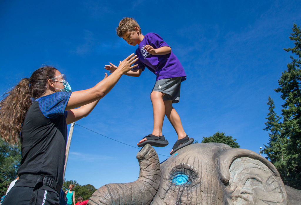 Haaken Williams, 6, balances on the nose of an elephant sculpture at Forest Park as Camp Prov unit leader Megan Pfohl reaches out during a playground break in Everett. (Andy Bronson / The Herald)
