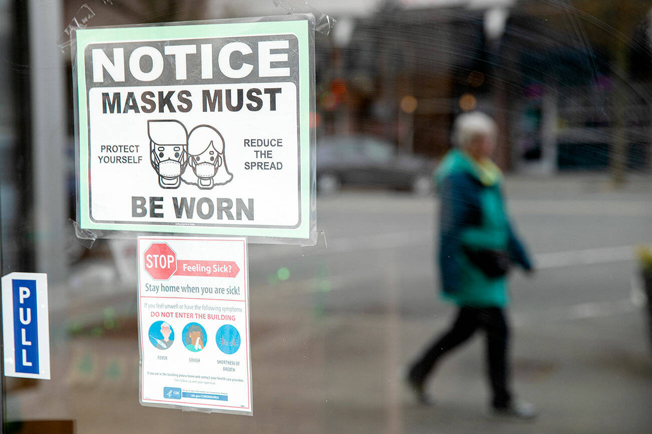 A woman walks past a downtown business with a sign requiring customers to wear masks Thursday, March 10, 2022, in Everett, Washington. (Ryan Berry / The Herald)
