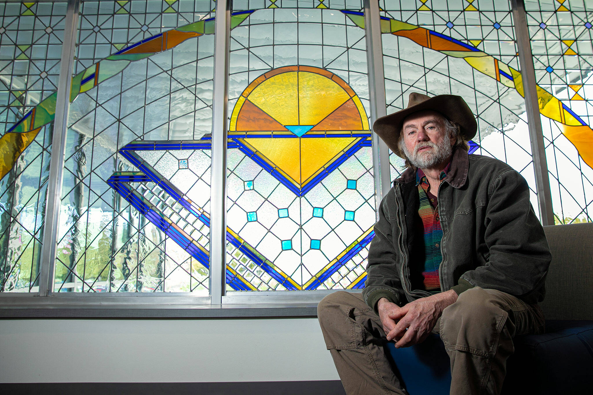 Jack Archibald, an artist who lives on Camano Island, sits in front of a stained glass window he created on the second floor of Stanwood High School. (Ryan Berry / The Herald)