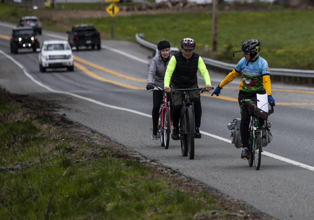 Kristin Kinnamon points to a spot that needs a Dan Henry directional mark as Steve Boskovich and Frances Philbin ride behind along Highway 532 on Sunday, April 10, 2022 in Camano Island, Washington. (Olivia Vanni / The Herald)
