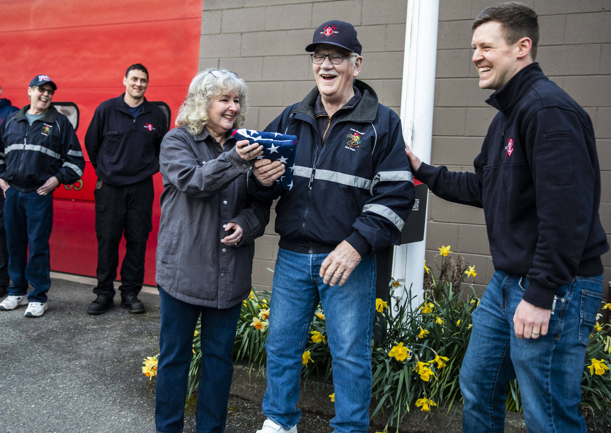 Marnie and Brian Anderson smile and laugh with Battalion Chief Justin Johnson (right) after receiving a flag as retirement gift at the Lake Roesiger Fire Department on Thursday in Snohomish. (Olivia Vanni / The Herald)