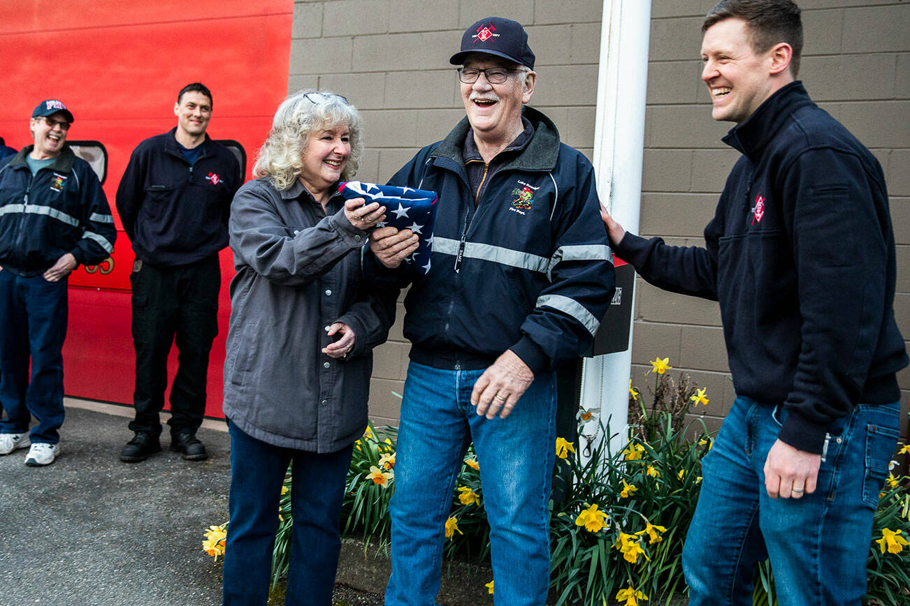 Marnie Anderson and Brian Anderson smile and laugh with Battalion Chief Justin Johnson, right, after receiving a flag as retirement gift at the Lake Roesiger Fire Department on Thursday, March 31, 2022 in Snohomish, Washington. (Olivia Vanni / The Herald)