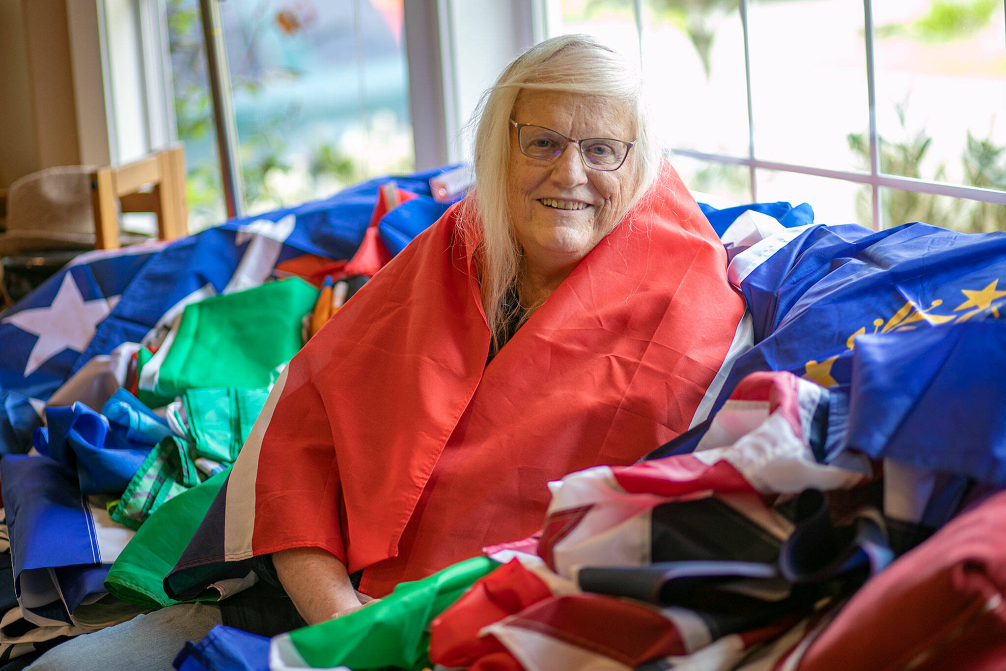 Melissa Batson, wrapped in the flag of The Gambia, sits among the 74 other flags she owns at her home in Monroe. Batson began flying different flags in front of her home in January 2021 to tell her journey as a transgender woman and to share her love of history, and has continued adding to her collection ever since. She posts photos on Facebook with biographical details or historical accounts. (Ryan Berry / The Herald)