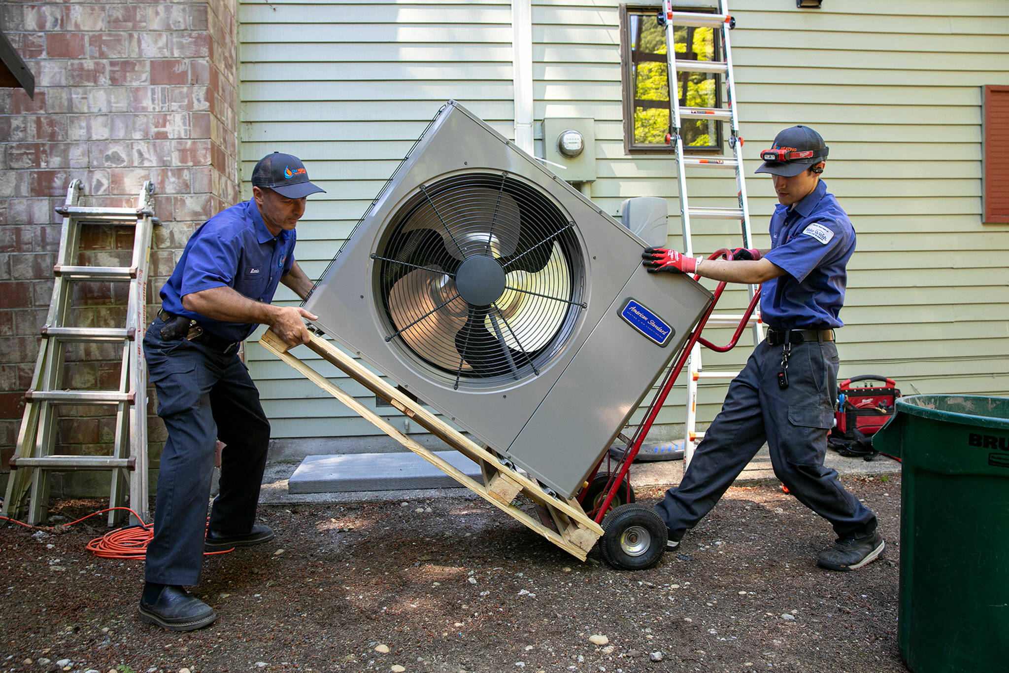 Remis Jankauskas, left, and Trent Pickford of CM Heating move a heat pump into place while working on a home’s HVAC system on Friday, in Woodinville. (Ryan Berry / The Herald)