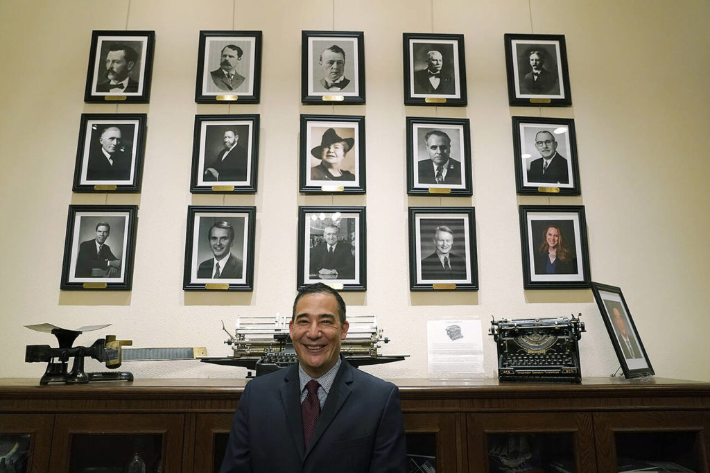 Steve Hobbs, who was sworn in as Washington Secretary of State, Monday, Nov. 22, 2021, at the Capitol in Olympia, poses in front of photos of the 15 people who previously held his office. (AP Photo/Ted S. Warren)
