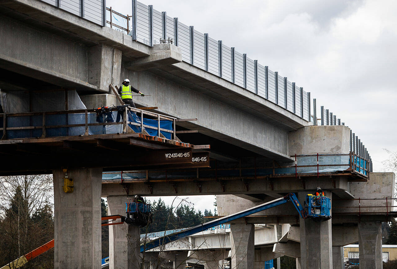 Construction workers work along the underside of the Lynnwood Link light rail guideway March 29 in Lynnwood. Delays caused by the pandemic and a concrete delivery strike earlier this year have led to a projected 6-month delay in the service’s start date. (Olivia Vanni / Herald file)
