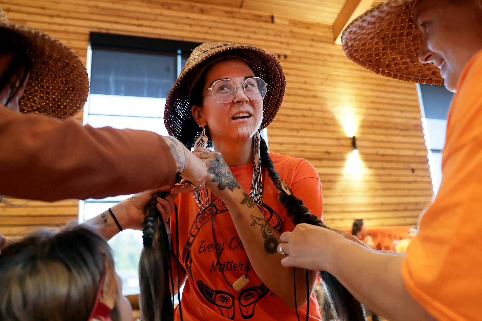 Kiera Blum, left, works to decorate Tayna Greene’s braids, center, with Linzie Crofoot, right, in preparation for a performance during the National Day for Truth and Reconciliation and Orange Shirt Day Friday evening at Tulalip Gathering Hall on Sept. 30, in Tulalip. (Kevin Clark / The Herald)