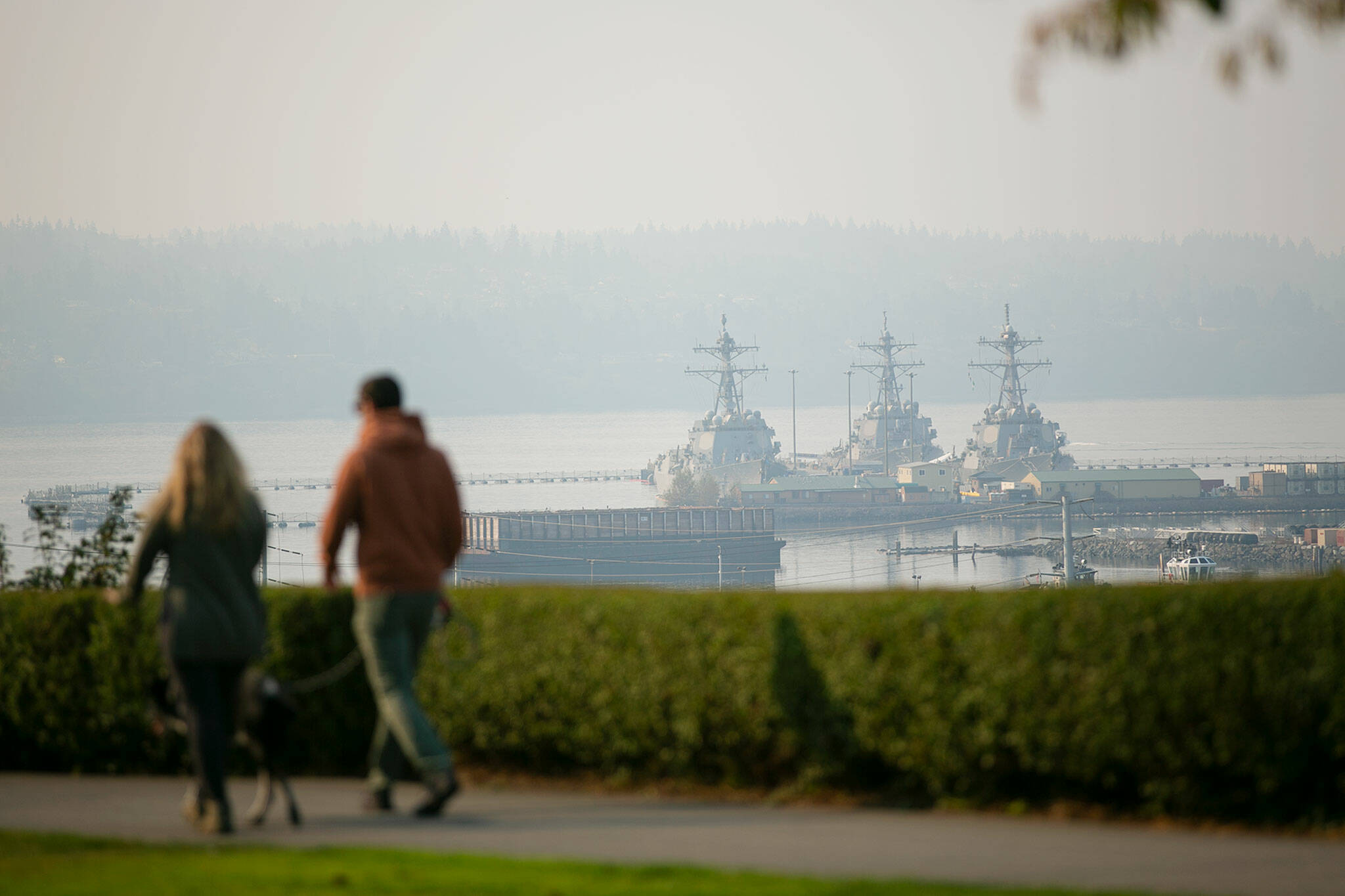 A couple walks their dog through Grand Avenue Park as smoke shrouds Naval Station Everett in the distance on Saturday, Oct. 15, 2022, in Everett, Washington. (Ryan Berry / The Herald)