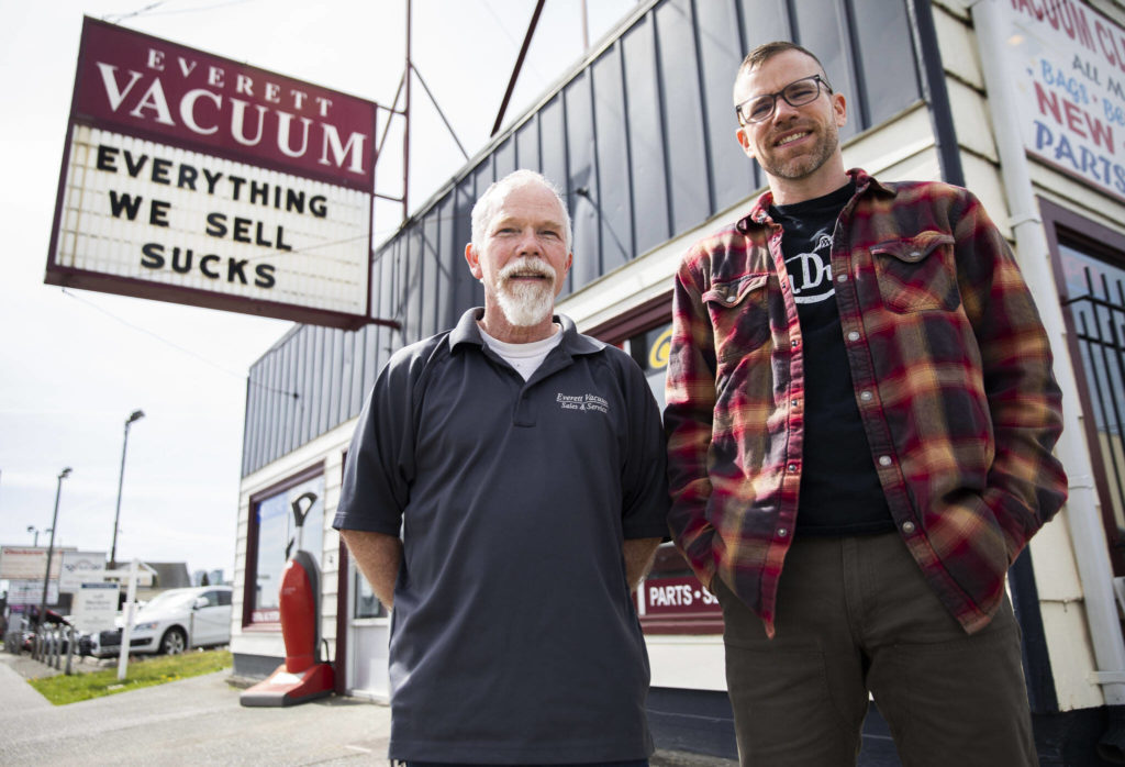 Father-and-son Mike and David Lane stand in front of Everett Vacuum on Broadway with their popular sign and saying, “Everything we sell sucks.” In November, the store moved to Hewitt Avenue, where overhang signs aren’t allowed, so a new flat banner with the sucks saying was made for the front window. (Olivia Vanni / The Herald)
