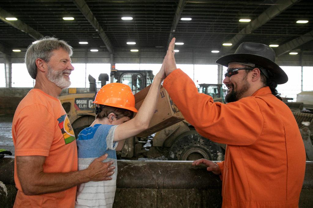 Joel Christensen, being supported by his father Craig, gives a round of high-fives to heavy equipment operator Shintaro Ishikawa, right, on Aug. 4, 2022, at the Airport Road Recycling and Transfer Station in Everett. Ishikawa and other employees at the station took notice of Joel’s frequent visits, and even gifted him a hardhat of his own as a way to recognize their appreciation for his presence. (Ryan Berry / The Herald)
