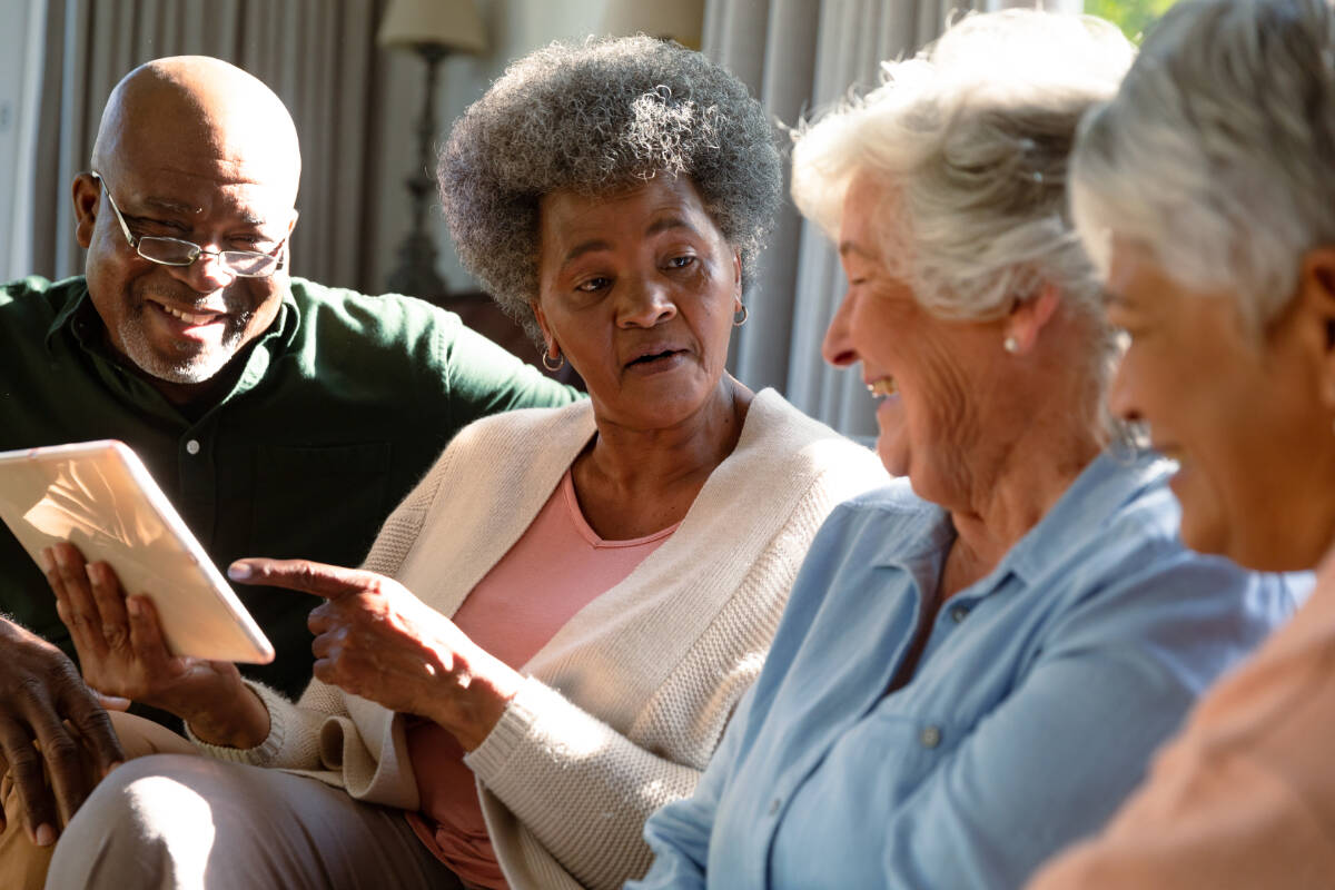 Three happy diverse senior woman and african american male friend sitting on sofa and using tablet. retirement lifestyle relaxing at home with technology.