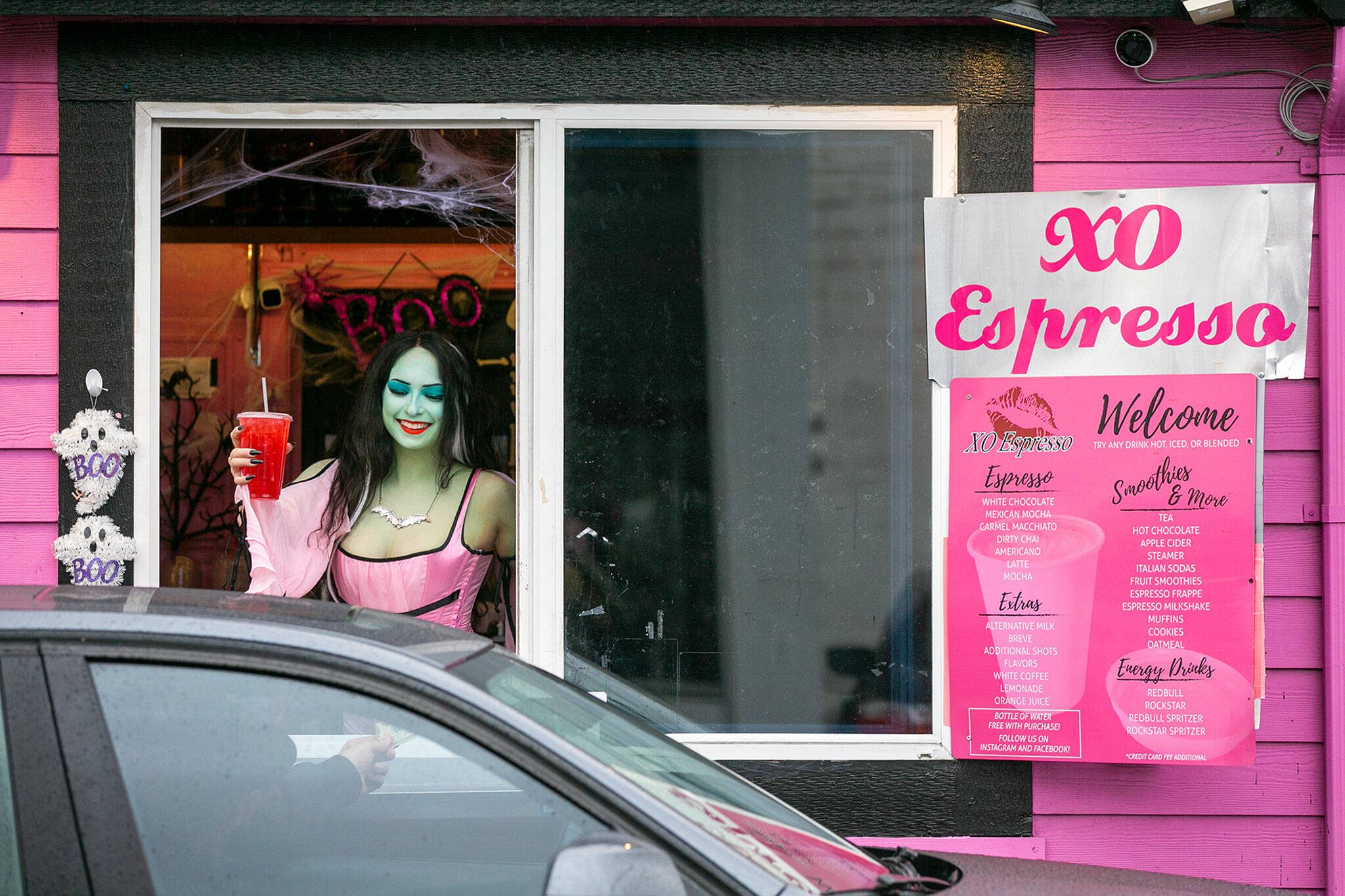 Emma Dilemma, a makeup artist and bikini barista for the last year and a half, serves a drink to a customer while dressed as Lily Munster Oct. 25, 2022, at XO Espresso on 41st Street in Everett, Washington. (Ryan Berry / The Herald)