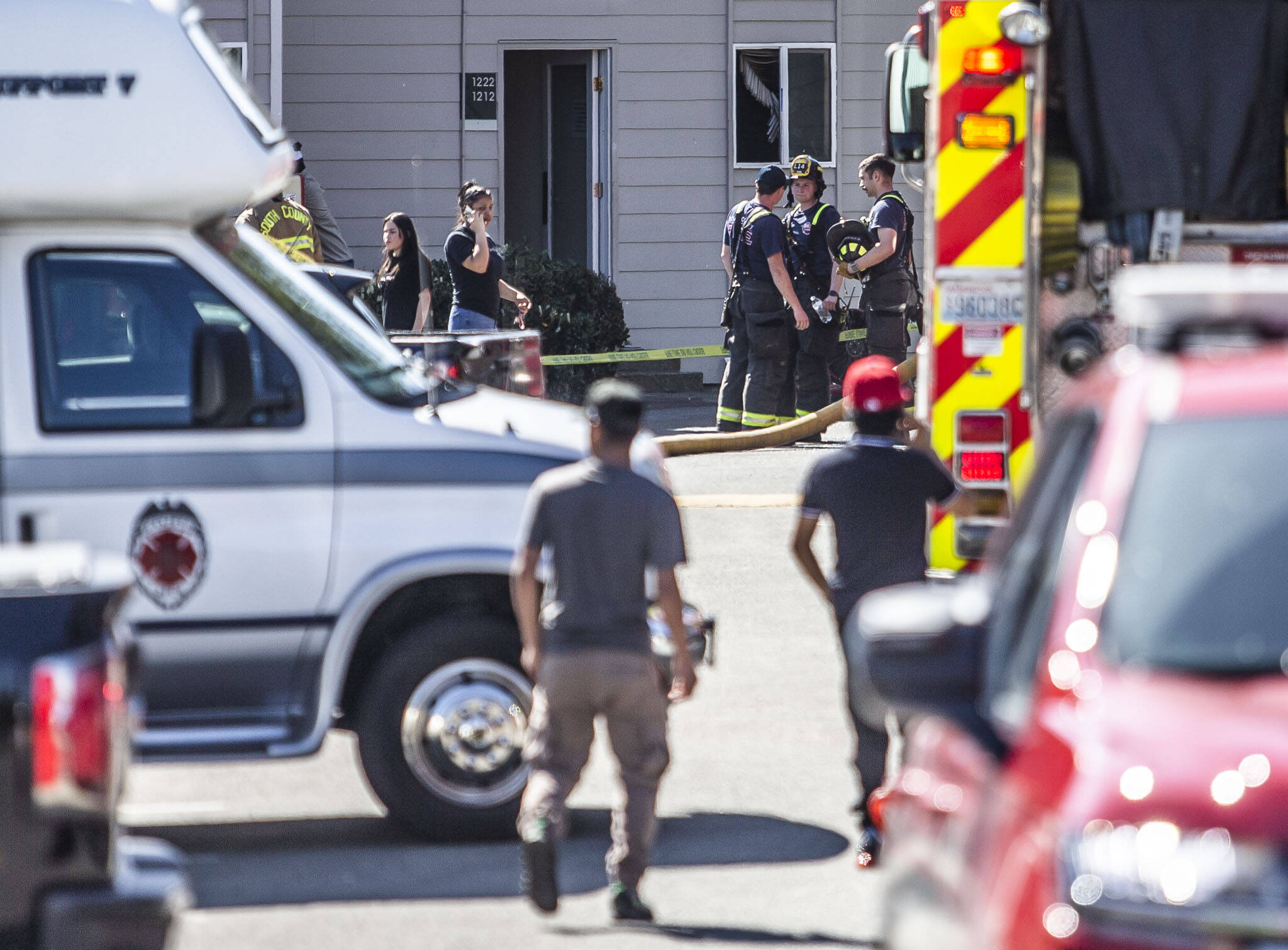 Firefighters and witnesses stand outside a building where damaged blinds are visible through the window of the scene of a fire on Thursday, May 11, 2023 in Lynnwood, Washington. (Olivia Vanni / The Herald)