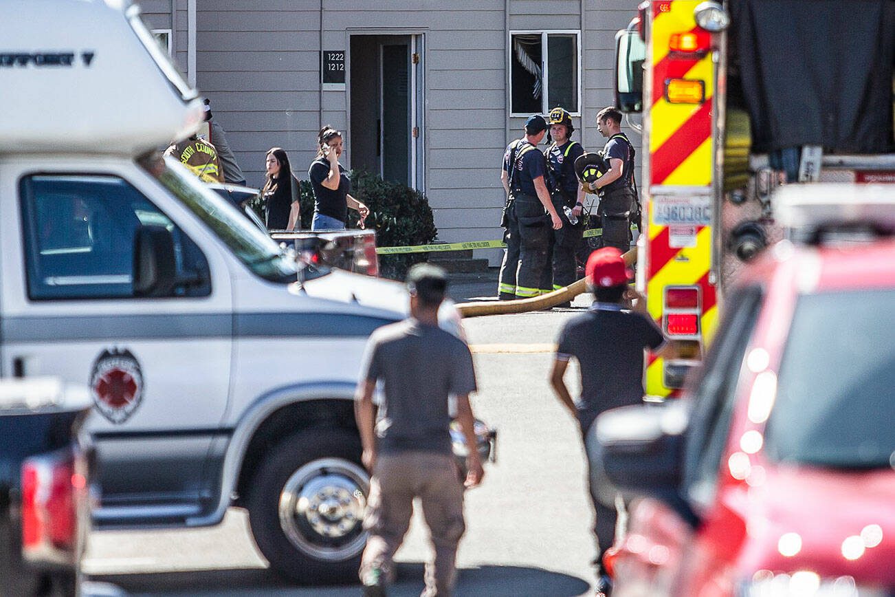 Firefighters and witnesses stand outside a building where damaged blinds are visible through the window of the scene of a fire on Thursday, May 11, 2023 in Lynnwood, Washington. (Olivia Vanni / The Herald)