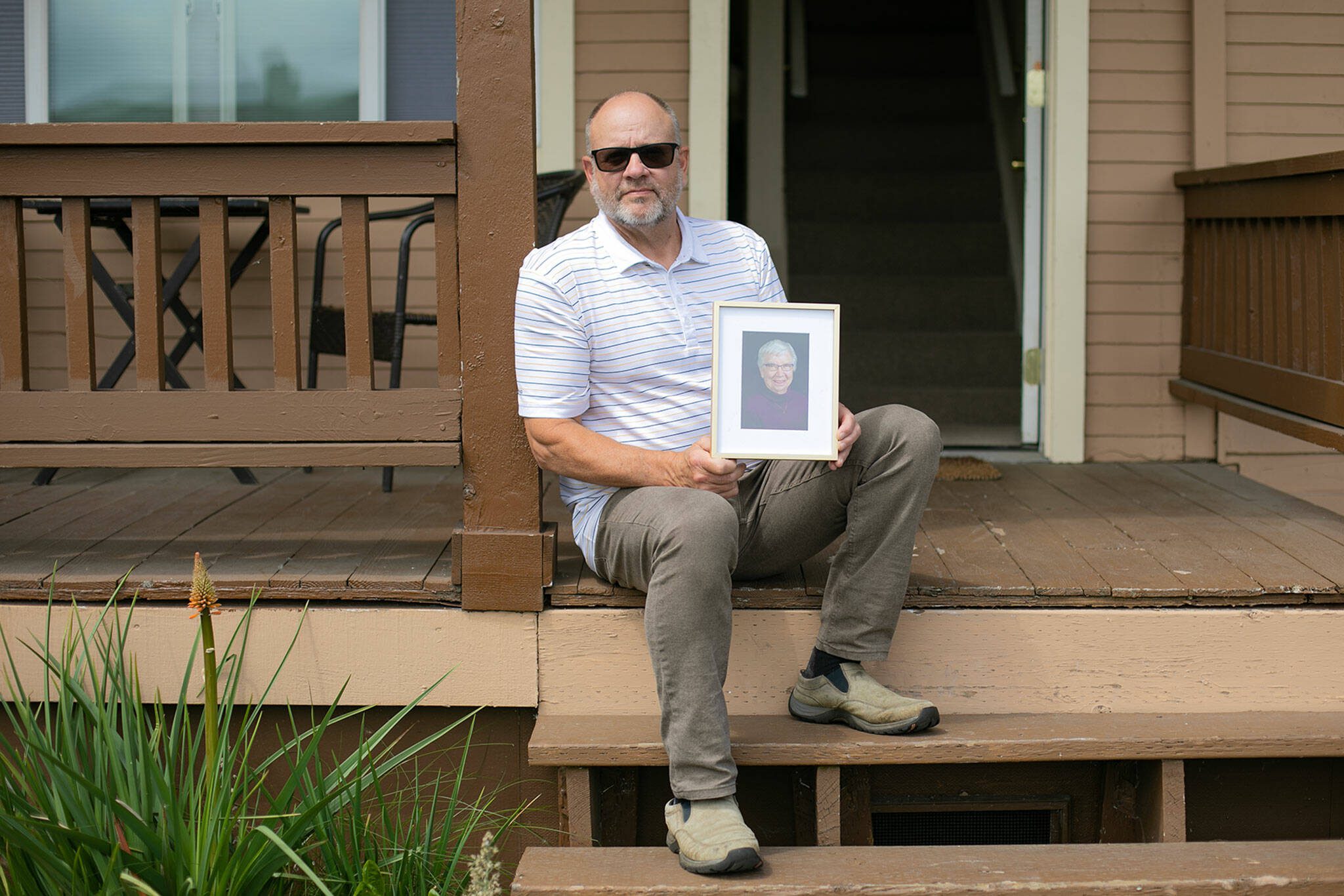 John Stejer sits in front of his home and holds a photo of his mother, Betty Stejer, 88, on Wednesday, May 24, 2023, in Everett, Washington. John moved his mother to Arizona in 2022 due to the difficulty of finding Medicaid-approved housing for her. (Ryan Berry / The Herald)