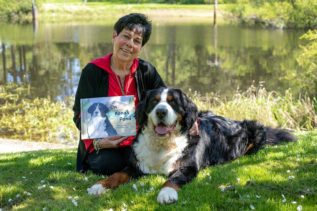 Michelle LeFevre and her Bernese mountain dog Kona sit in the shade in front of Kona’s Pond outside their home Wednesday, May 10, 2023, in Camano, Washington. LeFevre, a retired teacher, wrote the children’s book “On Kona’s Pond” which centers on her pup and the other creatures that call the pond home. LeFevre’s sister, Susan Cousineau McGough, illustrated the book with watercolor renditions of Kona and the pond. (Ryan Berry / The Herald)