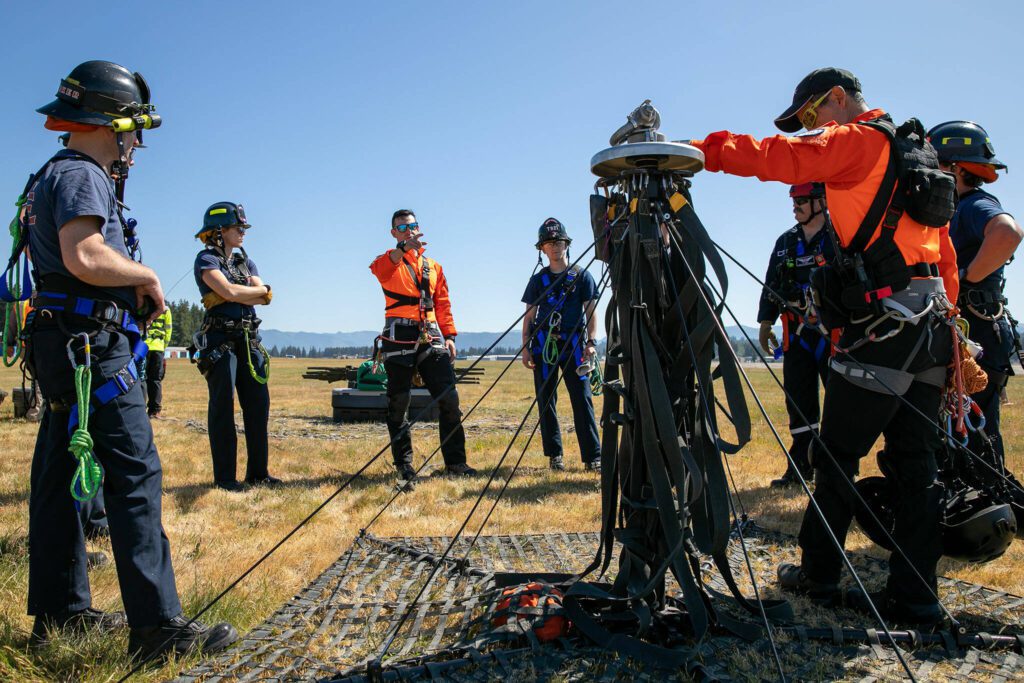 Snohomish County Volunteer Search and Rescue Flight Medic Jeff Brown, center left in orange, speaks with crews from South County Fire and Seattle Fire about the airborne tactical extraction platform (AirTEP) during an interagency disaster response training exercise at Arlington Municipal Airport on Tuesday, June 6, 2023, in Arlington, Washington. (Ryan Berry / The Herald)
