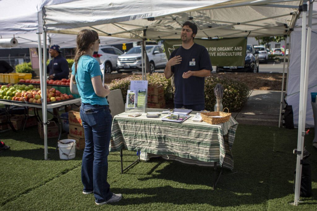 Joe Crumbley, right, speaks to a visitor at the Tilth Alliance booth at the Everett Farmers Market across from the Everett Station in Everett, Washington on Wednesday, June 14, 2023. (Annie Barker / The Herald)
