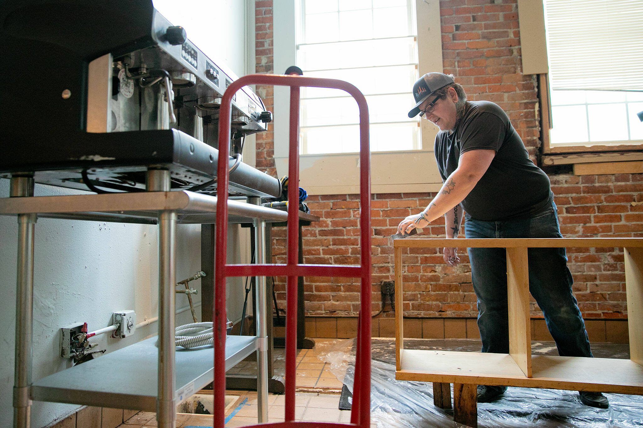 Adair Gearhart, working next to a 500-pound espresso machine, sands down a shelf they plan to repurpose while doing major renovations and cleaning at 1709 Hewitt Ave., the future downtown home of Catalyst Café, on Tuesday, June 20, 2023, in Everett, Washington. Gearhart said they hope to open later this year, though the massive scale of work to be done will ultimately dictate the opening date for the queer-friendly café. (Ryan Berry / The Herald)