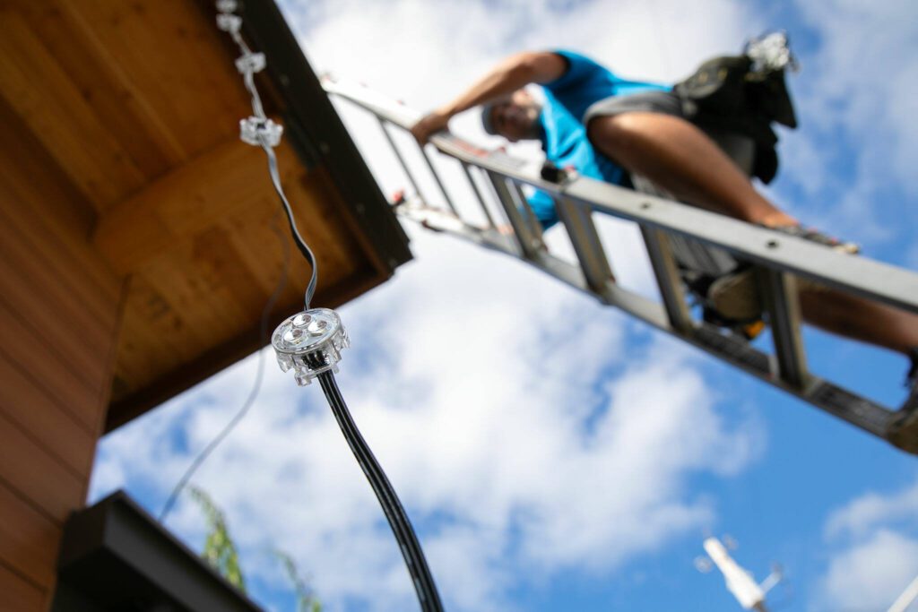 A strand of LED lights dangle from a new public restroom outside the Marysville Civic Center as Giles Electric owner Tyler Giles installs them on Friday, June 30, 2023, in Marysville, Washington. (Ryan Berry / The Herald)

