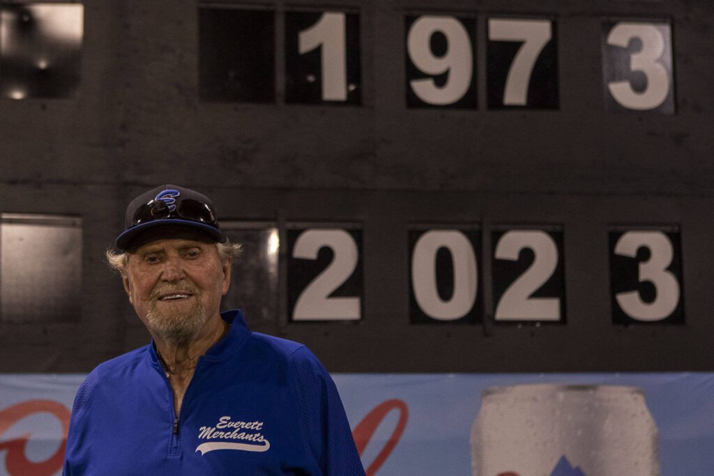 Everett Merchants manager Harold Pyatte poses next to a scoreboard displaying his years coaching after a game on Friday at Funko Field in Everett. (Annie Barker / The Herald)
