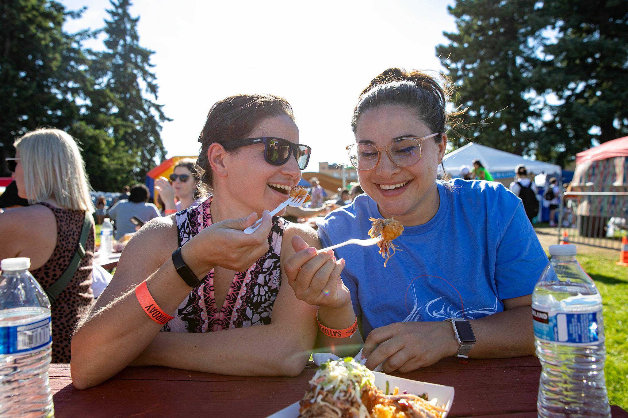 Sisters Nadia, left, and Yasmeen Kabbani dig into some pulled pork tater tots from Ryan’s REZ-ipes during Taste Edmonds on Saturday, August 12, 2023, at Franceós Anderson Play Field in Edmonds, Washington. (Ryan Berry / The Herald)