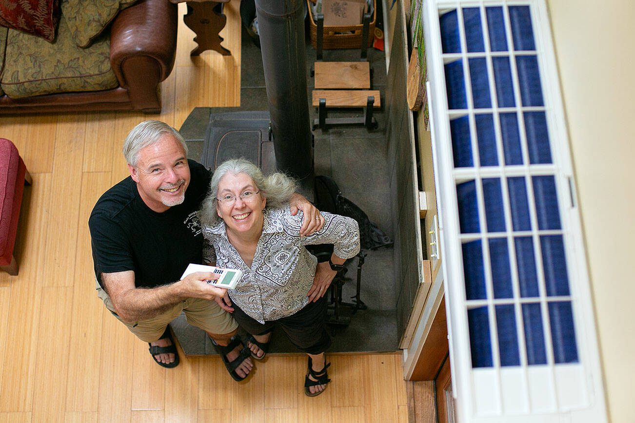 Homeowners Jim and Chris Hall stand beneath their new heat pump, at right, inside their Whidbey Island home on Thursday, Sep. 7, 2023, near Langley, Washington. The couple, who are from Alaska, have decreased their use of their wood burning stove to reduce their carbon footprint. (Ryan Berry / The Herald)