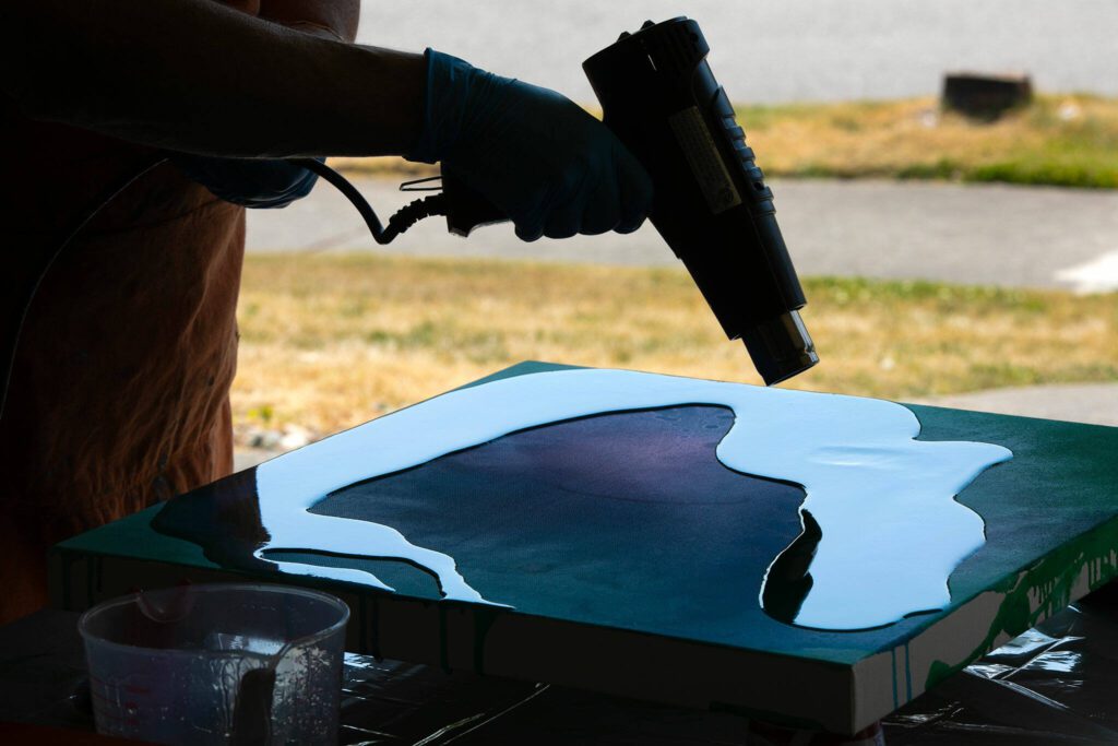 Downes uses a heating tool to remove bubbles from resin poured on a canvas. (Ryan Berry / The Herald)
