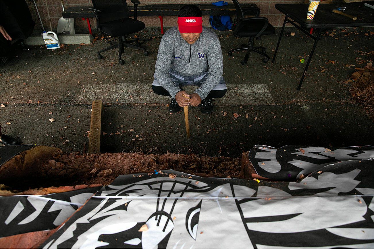 Master woodcarver James Madison squats next to the in-progress healing pole on Tuesday, June 13, 2023, at Archbishop Murphy High School in Everett, Washington. (Ryan Berry / The Herald)