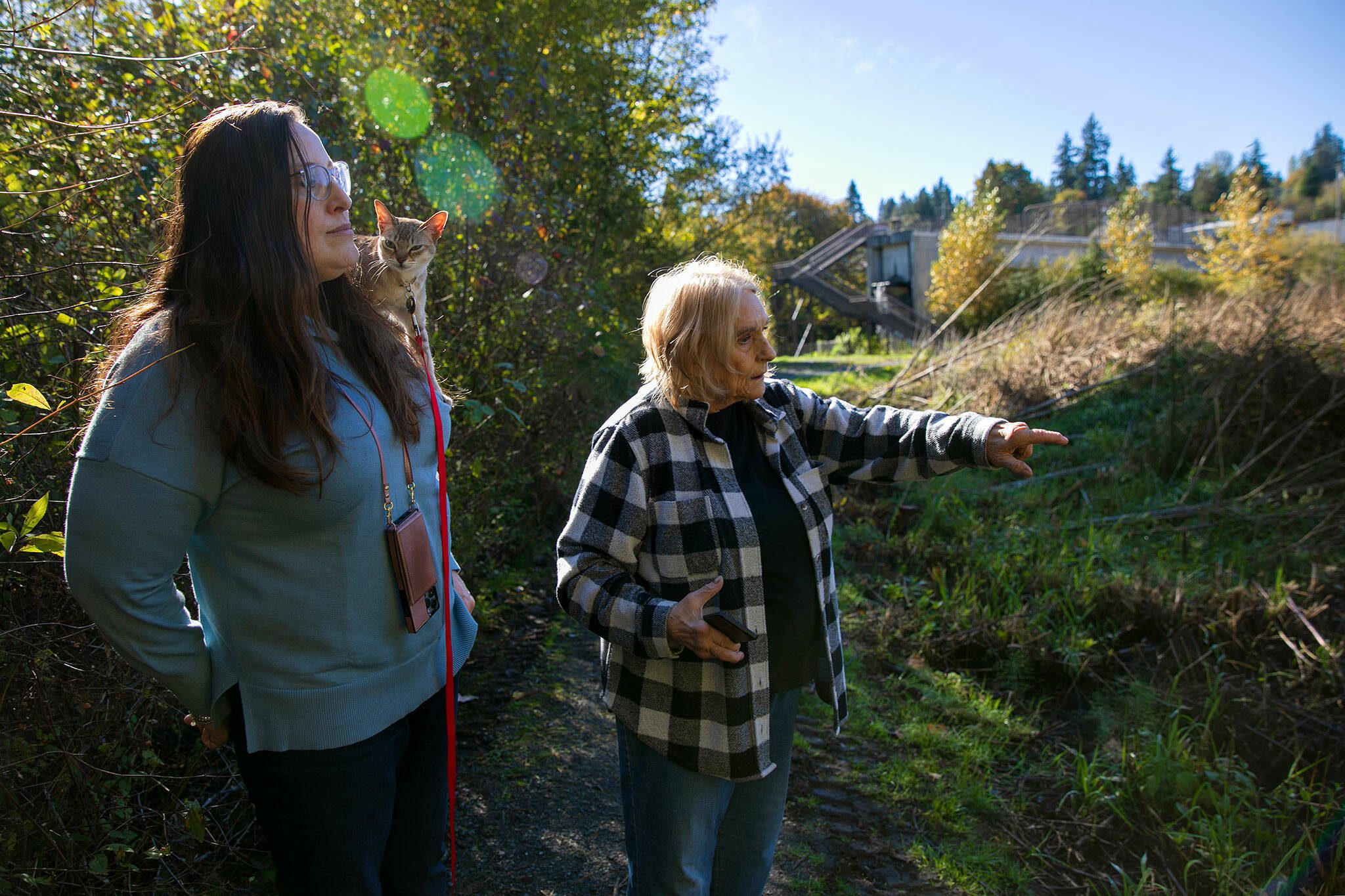 Brenda Bolanos-Ivory and her cat Piccolo, left, along with Gail Chism look over a section of cleared plant growth at Lowell Riverfront Park on Wednesday, Oct. 18, 2023, in Everett, Washington. The two Lowell residents feel the clearing of trees and undergrowth at the park is unacceptable. (Ryan Berry / The Herald)