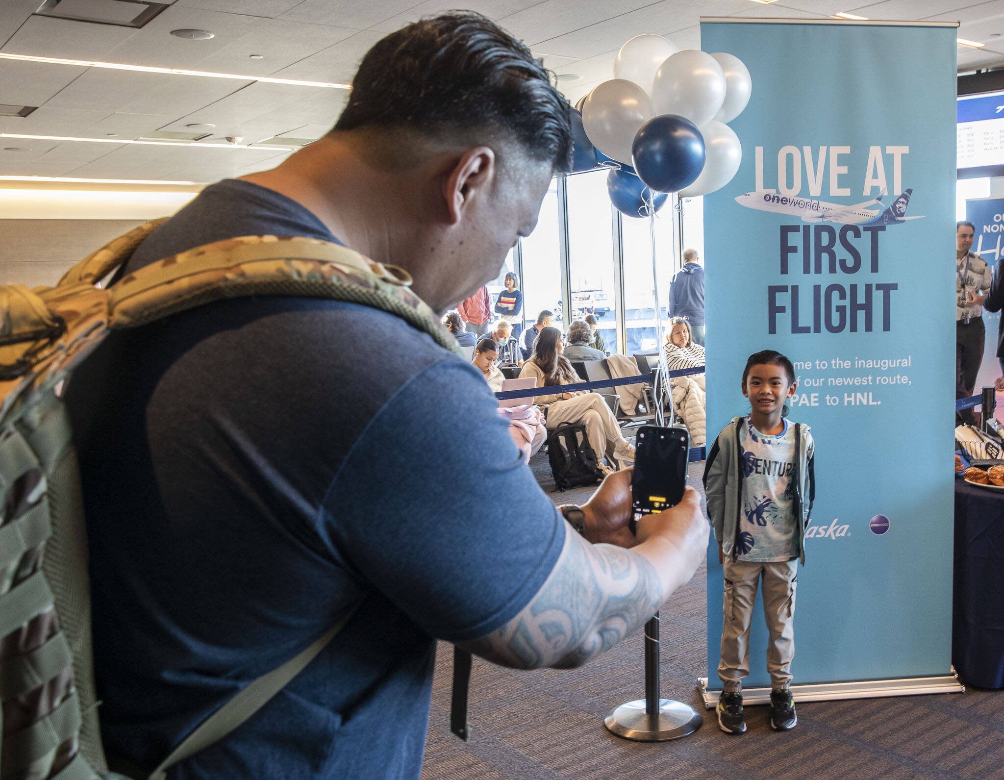 Ron Rendorio, left, takes a photo of Kamron, 6, in front of a sign announcing Alaska’s first flight to Honolulu from Paine Field on Friday, Nov. 17, 2023 in Everett, Washington. (Olivia Vanni / The Herald)