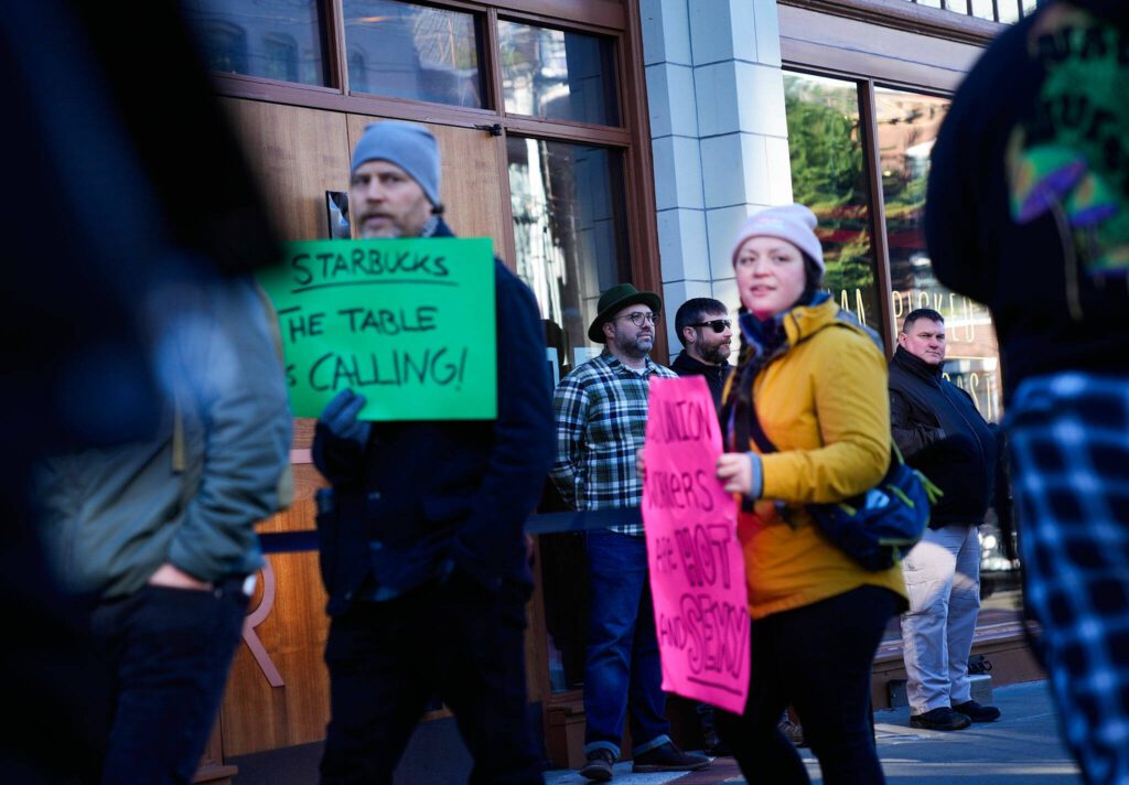 Starbucks manager Jeremy Jaureguito, in plaid, monitors the doors with security guards as workers participate in a walkout and strike organized by Starbucks Workers United during the company’s Red Cup Day Thursday, Nov. 16, 2023, at the company’s first Reserve roastery in Seattle. (AP Photo/Lindsey Wasson)
