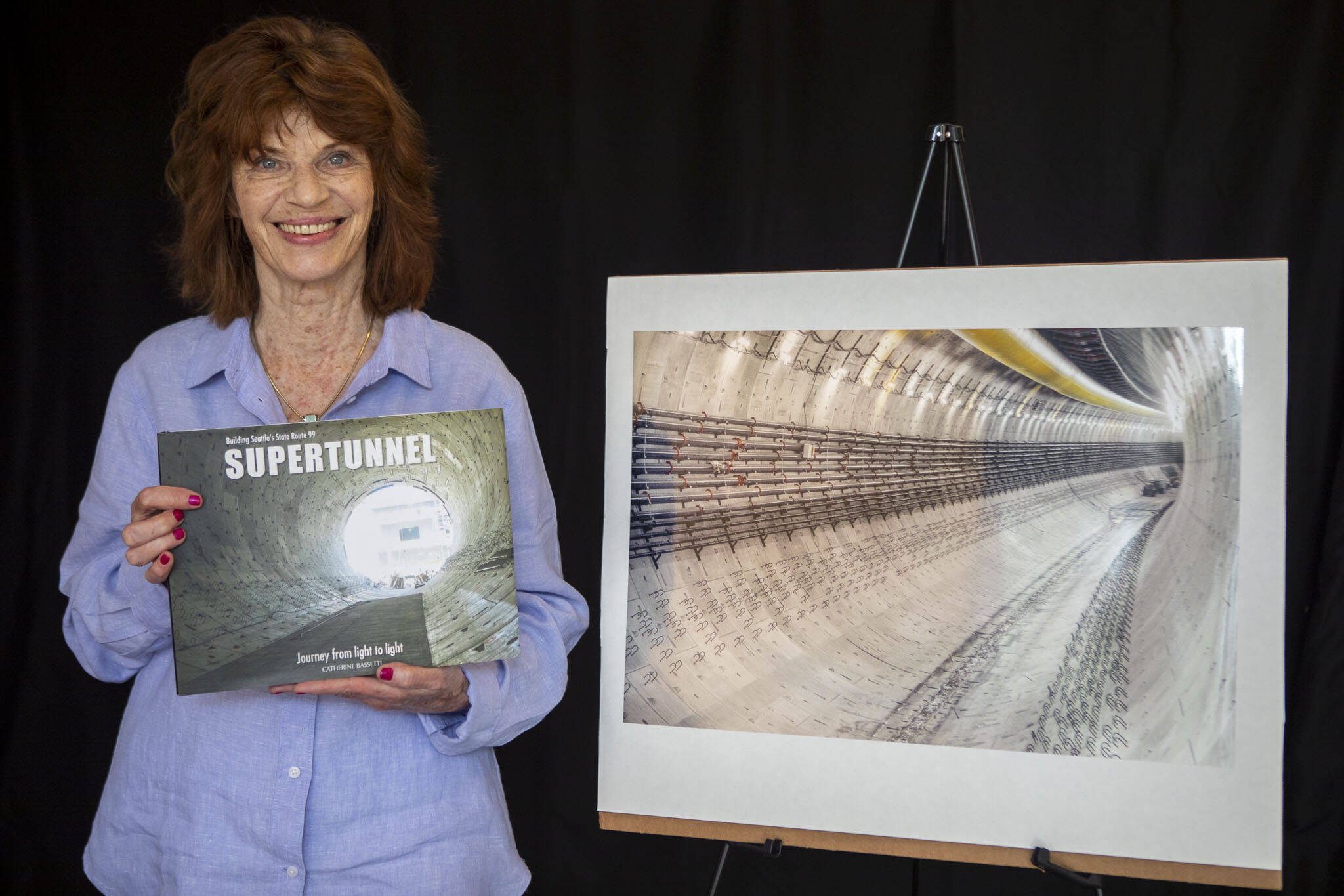 Catherine Bassetti poses for a photo with her book “Building Seattle’s State Route 99 Supertunnel: Journey from Light to Light” in her home studio in Shoreline, Washington on Friday, May 19, 2023. (Annie Barker / The Herald)