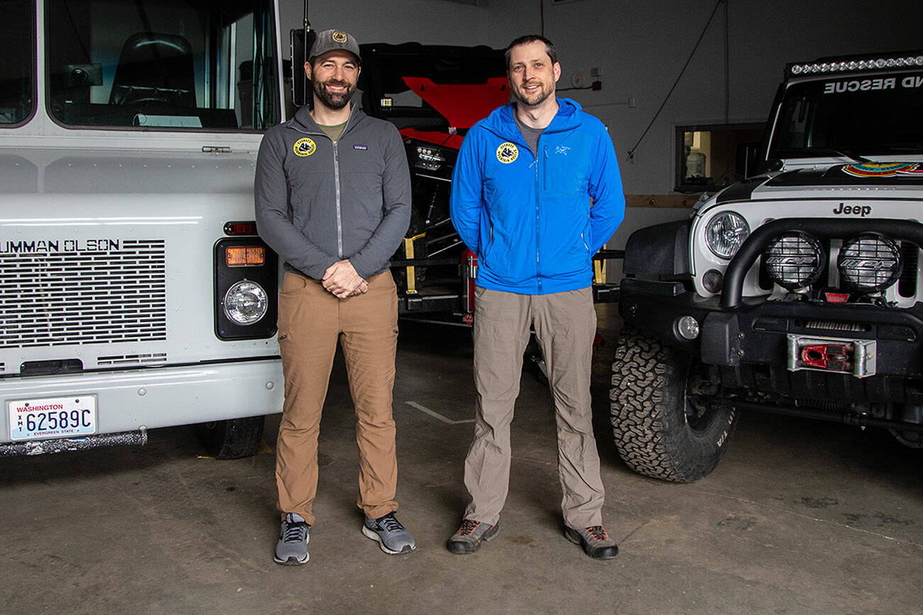 Field members Donnie Pingrey, left, and Jay Bublitz, right, pose for a photo in front of vehicles at the Everett Mountain Rescue headquarters in Snohomish, Washington on Thursday, Nov. 16, 2023. (Annie Barker / The Herald)