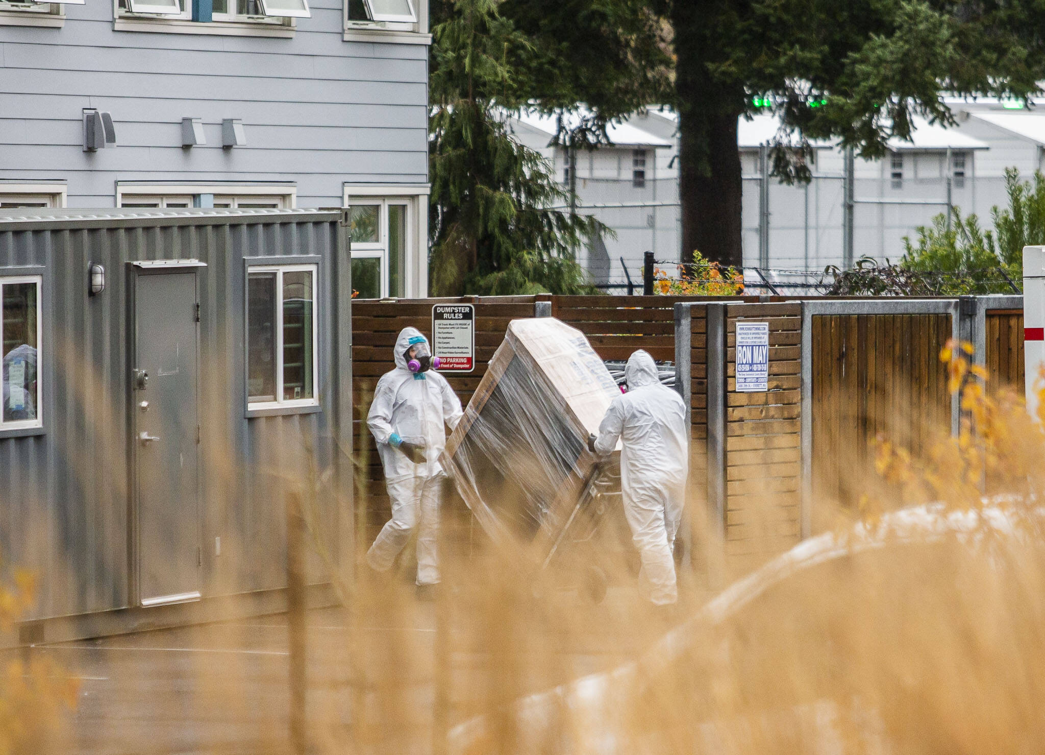 Two people in white protective suits move a large package out of Clare’s Place and into a storage container in the parking lot on Monday, Dec. 4, 2023 in Everett, Washington. (Olivia Vanni / The Herald)