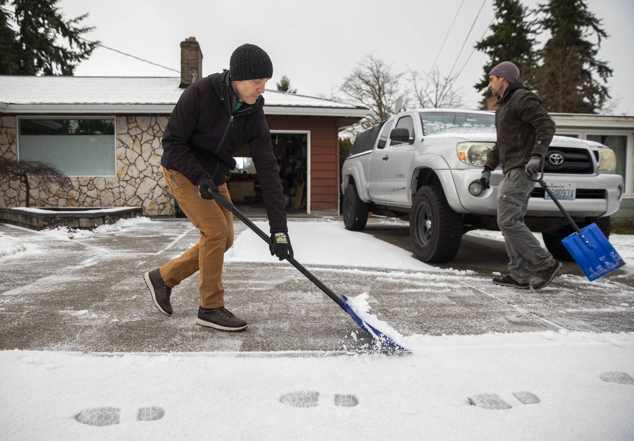 Shanti Curran, left, and Andy McKee, right, shovel Curran’s driveway after a light dusting of snow fell on Thursday morning in Everett, Washington. Curran said he normally wouldn’t shovel with such a small amount of snow but the looming below freezing temperatures over the next few days have him worried about potential ice. (Olivia Vanni / The Herald)