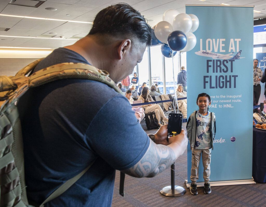 Ron Rendorio, left, takes a photo of Kamron, 6, in front of a sign announcing Alaska’s first flight to Honolulu from Paine Field on Nov. 17, 2023 in Everett. (Olivia Vanni / The Herald)
