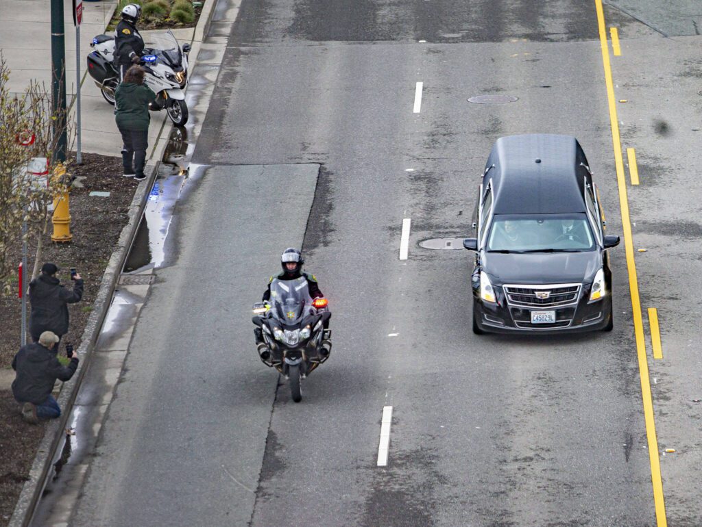 People stand along Marine View Drive and watch the hearse carrying Washington State trooper Christopher Gadd’s remains in a memorial procession on Tuesday, March 12, 2024, in Everett, Washington. (Olivia Vanni / The Herald)
