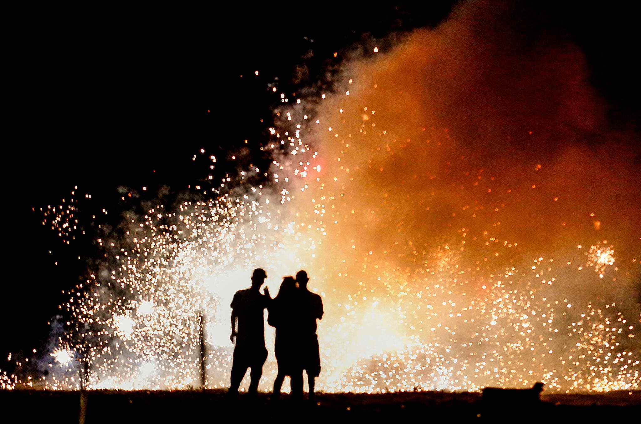 A trio in the lighting area at Boom City watches a shower of white-hot sparks from several fountains set off Tuesday just as darkness falls. (Dan Bates / The Herald)