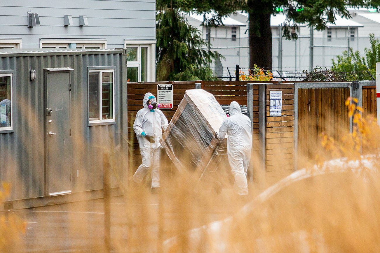 Two people in white protective suits move a large package out of Clare’s Place and into a storage container in the parking lot on Monday, Dec. 4, 2023 in Everett, Washington. (Olivia Vanni / The Herald)