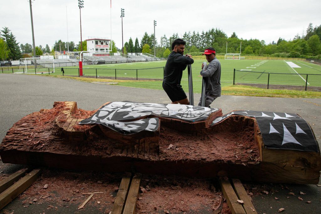 Archbishop Murphy High School facilities staffer Tani Tupou and artist James Madison, right, use metal poles to slide a 12-foot redwood healing pole back under cover on Tuesday, June 13, 2023, in Everett, Washington. (Ryan Berry / The Herald)
