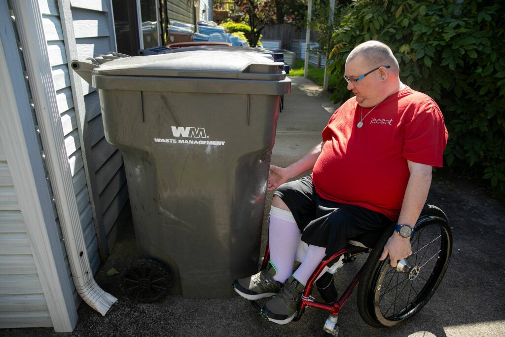 Ryan Carlson, 37, talks about how he takes garbage and recycling push carts to the curb while using his wheelchair at his group home on Friday, June 7, 2024, in Marysville, Washington. Carlson receives assistance from an aid to wrap a bungee cord around the container and then behind his legs in order to maintain control as he takes the bins to the curb. (Ryan Berry / The Herald)
